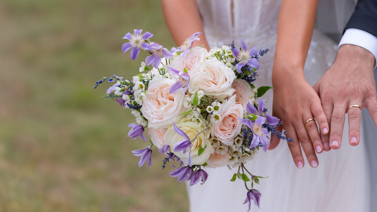 Bouquet da sposa con rose cipria e fiori lilla in primo piano, tra le mani degli sposi